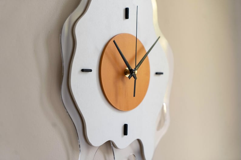May include: A quirky wall clock shaped like a fried egg. It has a white, irregular border, a light brown central circle, and black clock hands. The clock's dimensions are not visible in the image.