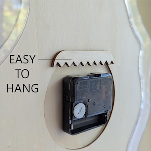 May include: Close-up of the back of a clock, with a black clock mechanism and a wooden sawtooth hanger. The text "EASY TO HANG" is printed on the light-coloured wood surface. The clock has a clear, curved edge.