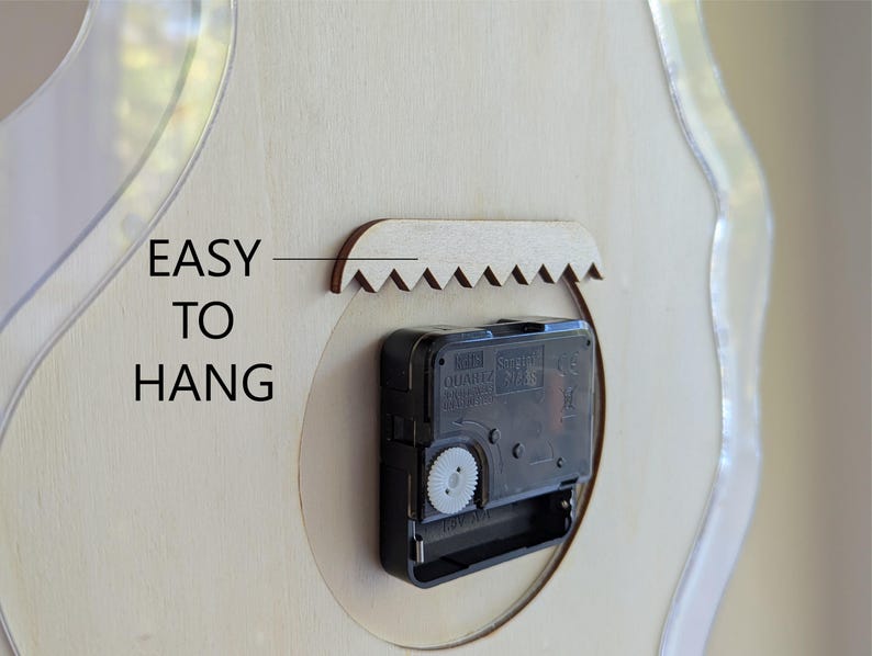 May include: Close-up of the back of a clock, with a black clock mechanism and a wooden sawtooth hanger. The text "EASY TO HANG" is printed on the light-coloured wood surface. The clock has a clear, curved edge.