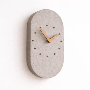 Puede incluir: Un reloj de pared gris de forma ovalada con manecillas de madera y pequeños puntos oscuros que marcan las horas. El reloj tiene una superficie texturizada y un diseño minimalista, adecuado para la decoración del hogar moderna.