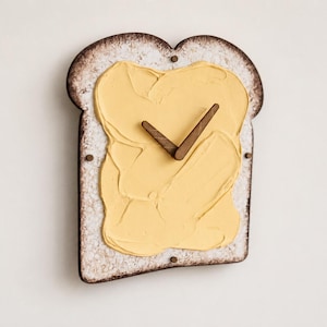 Op de afbeelding: Een wandklok in de vorm van een snee geroosterd brood met een textuur van lichtgele boter in het midden. De klok heeft donkerbruine houten wijzers en kleine donkerbruine ronde markeringen aan de boven-, linker- en rechterkant.