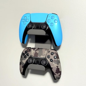 May include: Two game controllers, one light blue and one camouflage, mounted on a gray wall holder. The blue controller has black accents, while the camouflage controller features a mix of brown, gray, and black patterns. The holder is designed to display the controllers.