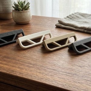 May include: Four textured cabinet pulls in black, off-white, tan, and grey are arranged on a wooden surface. Each pull has a geometric design with an open handle. A small succulent plant in a ceramic pot is in the background.