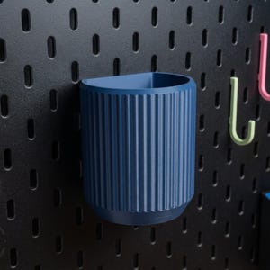 Peut inclure: Un gobelet de rangement cylindrique bleu marine, nervuré, est monté sur un panneau perforé noir. Deux crochets, un rose et un vert, sont également fixés. Une petite étagère de couleur claire avec une boîte est visible.