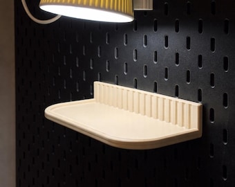 May include: A cream-colored shelf with a ribbed back panel is mounted on a black pegboard wall. A yellow lampshade hangs above the shelf, casting light. The shelf is designed for storage or display.