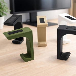 Può includere: Una collezione di supporti per cuffie moderni e minimalisti in vari colori: nero, bianco, verde oliva e beige. Ogni supporto presenta una parte superiore rettangolare con una superficie strutturata e una base curva a forma di gancio.