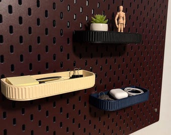 May include: Three rectangular wall-mounted storage trays with ribbed sides in black, cream, and dark blue, attached to a dark maroon pegboard. The trays hold small items including a succulent, a wooden mannequin, sticky notes, and keys.