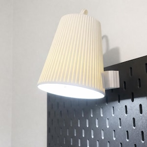 May include: A cream-colored, cone-shaped lamp with vertical ridges is mounted on a black pegboard. The lamp is lit, casting a warm glow. The lamp is attached to a white bracket.