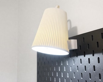 May include: A cream-colored, cone-shaped lamp with vertical ridges is mounted on a black pegboard. The lamp is lit, casting a warm glow. The lamp is attached to a white bracket.