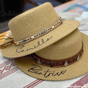 Può includere: Due cappelli di paglia con bande decorative. Il cappello superiore ha un fiocco di pelle marrone e il testo "Camilla" scritto su di esso. Il cappello inferiore ha una fascia di pelle marrone con accenti metallici e il testo "Beatrice" scritto su di esso.