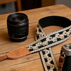 Può includere: Una tracolla per fotocamera nera con un design decorativo con perle e finiture dorate. La tracolla ha un'estremità in pelle marrone con il marchio "Beatrice" stampato sopra. Sono visibili anche un obiettivo e un corpo macchina.