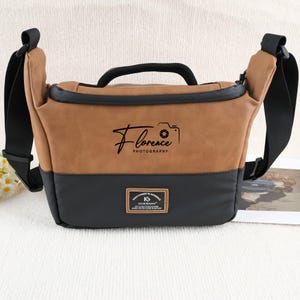 Könnte beinhalten: Eine braune und schwarze Kameratasche mit schwarzem Griff und Schultergurt. Auf der Tasche steht "Florence Photography". Am unteren Rand der Tasche befindet sich ein kleines rechteckiges Etikett.