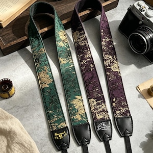 May include: Two camera straps featuring a textured pattern with gold accents. One strap is dark green, and the other is deep purple. Both have black leather ends, with the name Hannah printed on the green strap and Beatrice Photography on the purple one.