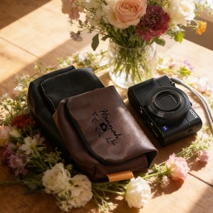 Könnte beinhalten: Eine schwarze Digitalkamera mit einem Zeiss-Objektiv liegt neben zwei Kamerataschen aus Leder, eine schwarze und eine braune. Die braune Tasche ist mit dem Namen "Hannah" und einem Kamera-Symbol personalisiert. Ein Blumenstrauß ist im Hintergrund.