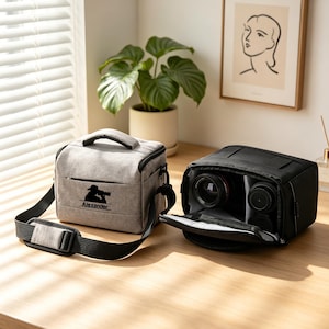 Pode incluir: Uma bolsa de câmara cinzenta clara com uma alça preta e um gráfico preto de um fotógrafo com o nome Alexander impresso por baixo, ao lado de uma bolsa preta aberta contendo duas lentes com elementos de vidro visíveis.
