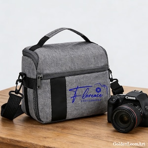 May include: Gray camera bag with a black handle and shoulder strap. The bag has a black stripe down the front and the text "Florence Photography" in blue. A Canon camera is next to the bag.