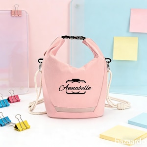 Könnte beinhalten: Eine hellrosa Lunchtasche mit schwarzem Verschluss und weißem Schultergurt. Die Tasche trägt den Namen "Annabelle" in Schwarz, innerhalb eines Kamera-Symbols. Die Tasche besteht aus weichem, wasserdichtem Material.