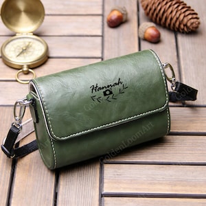 Può includere: Borsa per fotocamera in pelle verde con il nome "Hannah" e un'icona della fotocamera. La borsa ha una tracolla nera e finiture argentate. La borsa è rettangolare con chiusura a patta e cuciture bianche.