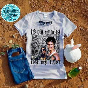 May include: A white t-shirt with a graphic featuring two men and the text "IT'S NOT my FIRST OR my LASTS". The shirt has a pair of sunglasses on it. A pair of blue jeans and a white rabbit are next to the shirt.