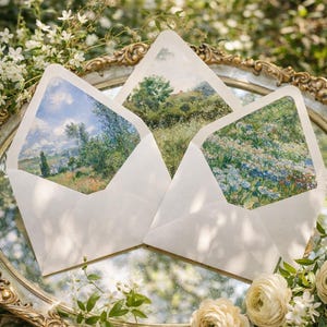 以下が含まれることがあります： 装飾的な鏡の上に展示された、風景画が内側に入った3つの白い封筒。鏡は白い花と緑に囲まれています。封筒には、木、畑、青い空のあるさまざまな牧歌的なシーンが描かれています。