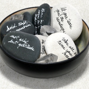 May include: A black bowl filled with smooth, gray and white stones. Each stone has a handwritten message in white ink. The messages include "let your tears join the raindrops", "take all the time you need", "hold tight memories", "not grief but gratitude", "there is more room in a broken heart", and "it's okay".