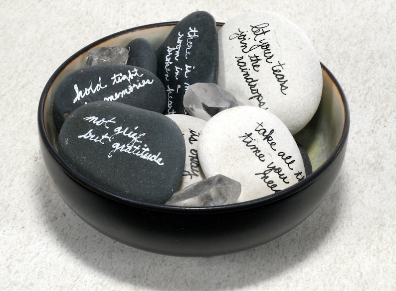 May include: A black bowl filled with smooth, gray and white stones. Each stone has a handwritten message in white ink. The messages include "let your tears join the raindrops", "take all the time you need", "hold tight memories", "not grief but gratitude", "there is more room in a broken heart", and "it's okay".