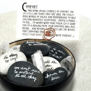 May include: A black bowl filled with smooth, gray and white stones. Each stone has a handwritten message of comfort and encouragement. The bowl also contains clear quartz crystals. The text on the card reads: "This bowl molds symbols of comfort. The crystals are tears that are shed. The pebbles hold words of solace and remembrance to help you find heartfelt acceptance. Choose a pebble... to feel... to warm with your touch. Let it guide you to a healing path. Your emotions will rise... recede... and return again just like the waves that smoothed these pebbles. Trust your heart. Be gentle with yourself."