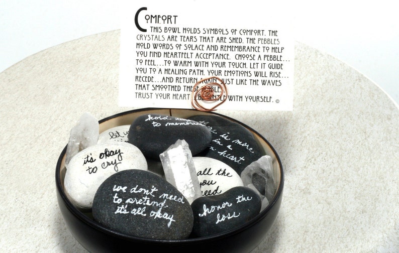 May include: A black bowl filled with smooth, gray and white stones. Each stone has a handwritten message of comfort and encouragement. The bowl also contains clear quartz crystals. The text on the card reads: "This bowl molds symbols of comfort. The crystals are tears that are shed. The pebbles hold words of solace and remembrance to help you find heartfelt acceptance. Choose a pebble... to feel... to warm with your touch. Let it guide you to a healing path. Your emotions will rise... recede... and return again just like the waves that smoothed these pebbles. Trust your heart. Be gentle with yourself."