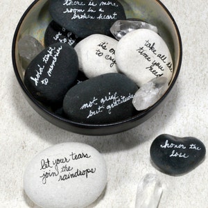 May include: A bowl filled with smooth, gray and white stones, each with an inspirational message written in white ink. The bowl is on a white surface, and there are two additional stones and a clear crystal on the surface. The stones read: "there is more room in a broken heart", "hold tight to memories", "it's ok to cry", "take all the time you need", "not grief but gratitude", "let your tears join the raindrops", and "honor the loss".