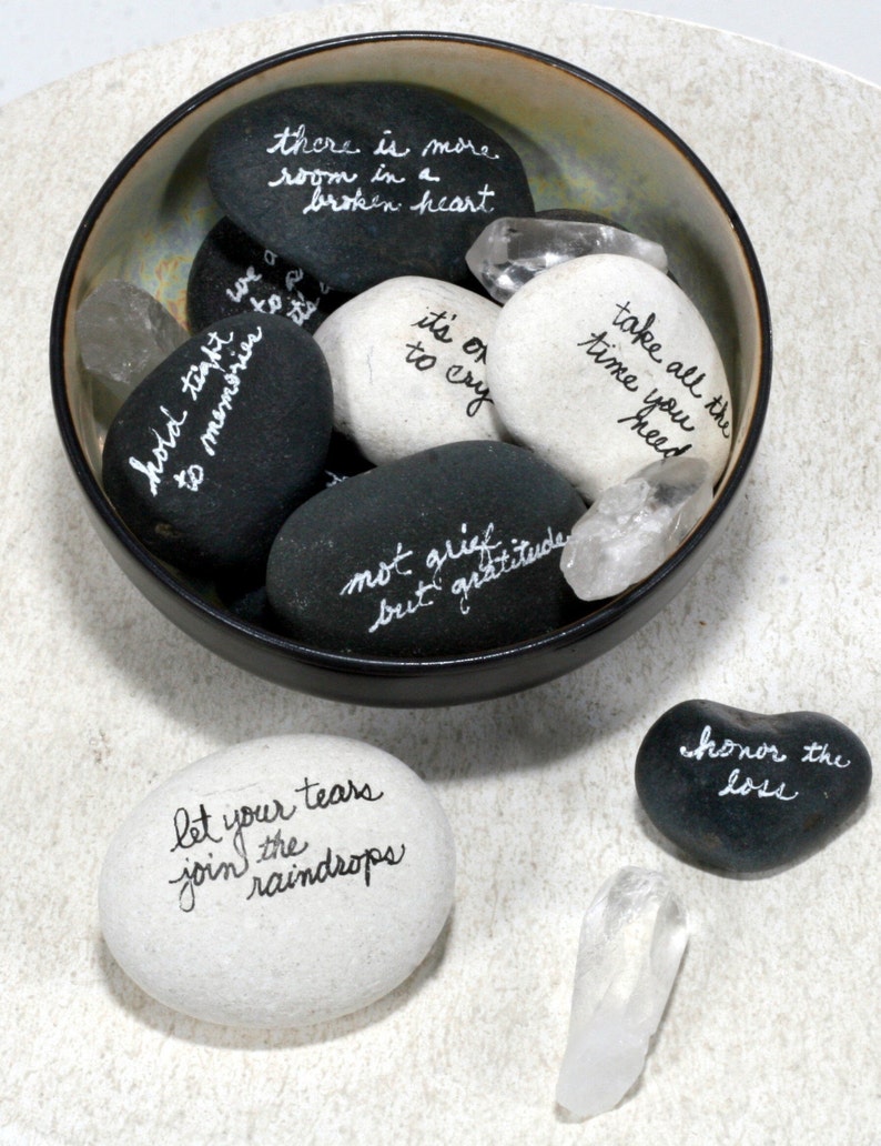 May include: A bowl filled with smooth, gray and white stones, each with an inspirational message written in white ink. The bowl is on a white surface, and there are two additional stones and a clear crystal on the surface. The stones read: "there is more room in a broken heart", "hold tight to memories", "it's ok to cry", "take all the time you need", "not grief but gratitude", "let your tears join the raindrops", and "honor the loss".