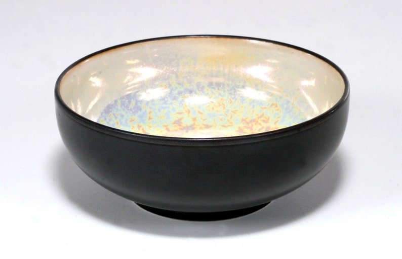 May include: A black ceramic bowl with a white iridescent interior. The bowl has a rounded shape and a small foot.