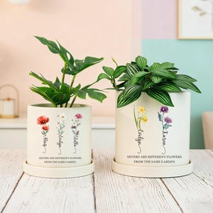 May include: Two off-white ceramic plant pots with plants. The pots feature floral designs and the names "Mag", "Evelyn", "Stella", and "Karen". The text "Sisters are different flowers from the same garden" is printed on the pots.