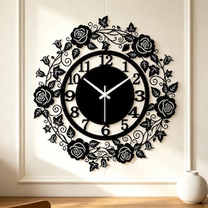 Könnte beinhalten: Eine runde Wanduhr mit schwarzem Zifferblatt, weißen Ziffern von eins bis zwölf und weißen Zeigern. Die Uhr ist von einem dekorativen schwarzen Metallrahmen umgeben, der wie ein Kranz aus Rosen, Blättern und Ranken geformt ist.