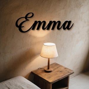 May include: A black metal sign in cursive script spelling "Emma" on a textured beige wall. A small wooden bedside table with a lamp is positioned below the sign.