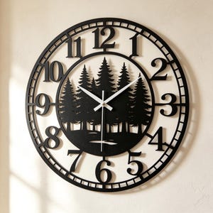 May include: A circular black wall clock featuring a cutout design of a forest with pine trees in the center. The clock face displays large black Arabic numerals from one to twelve arranged around the perimeter, with thin white hour and minute hands.