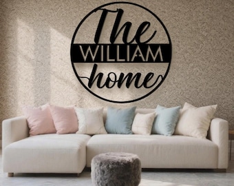 May include: A black metal sign reading "The WILLIAM home" in a circular design. It's mounted on a textured wall above a white sofa with pink, blue, and white cushions. A grey ottoman is in front of the sofa, creating a cozy living space.