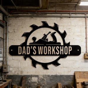 Puede incluir: Un letrero de madera negro con el texto "DAD'S WORKSHOP" en un taller. El letrero presenta un diseño de hoja de sierra con una silueta de cepillo en el centro. El letrero está montado en una pared de ladrillo blanco.