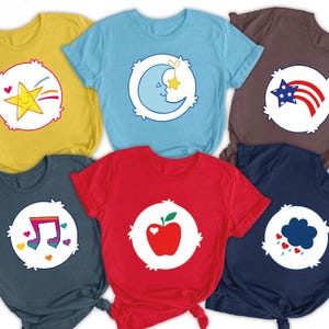 May include: A collection of colorful t-shirts featuring cartoon designs. Each shirt has a unique graphic on the front, including a star with a rainbow, a crescent moon, a patriotic star and stripes, musical notes, a red apple, and a cloud with hearts.