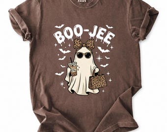 May include: A brown short-sleeve t-shirt featuring a graphic of a ghost wearing sunglasses, a leopard print bow, and holding an iced coffee and a leopard print handbag. The text BOO-JEE is printed above the ghost, surrounded by small bats and stars.