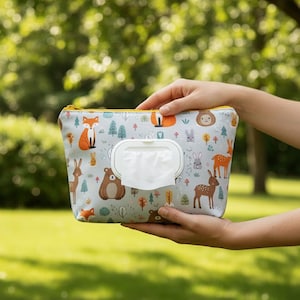以下が含まれることがあります： 黄色いジッパーと白いディスペンサーが付いた、明るい灰色のベビーワイプケース。ケースは、キツネ、鹿、クマなどの森の動物のパターンで装飾されています。ケースは、ぼやけた緑色の背景の前に保持されています。