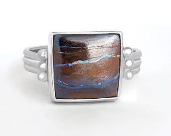May include: A square-faced ring with a silver band and a large, rectangular gemstone. The gemstone features a mix of brown, blue, and white colors. The ring has a unique design with small silver accents on the sides.