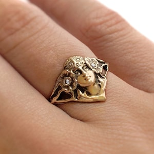 May include: A gold-toned metal ring featuring a sculpted face of a person wearing a decorative headpiece, flanked by two floral motifs. Each flower contains a small, clear, round inset stone. The ring has an oxidised finish in the recessed areas.