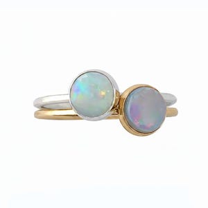Puede incluir: Dos anillos apilados con ópalos cabujón redondos engastados en bisel. Un anillo está hecho de plata pulida con un ópalo blanco que muestra destellos iridiscentes, mientras que el otro es de oro con un ópalo translúcido de tonos sutiles.