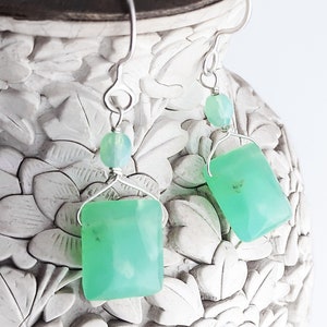 May include: A pair of silver earrings with green gemstone square drops and smaller round green beads. The earrings are hanging from a white ceramic vase with floral designs.