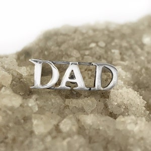 May include: Silver charm with the word "DAD" in block letters.