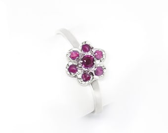 May include: A silver ring featuring a flower design. The flower is composed of seven round, deep pink gemstones, with a larger gemstone at the center. The ring is set against a white background.