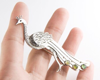 Puede incluir: Broche de pavo real plateado con un diseño detallado. El broche presenta un pavo real con corona, plumas texturizadas y una larga cola. La cola tiene tres gemas verdes y amarillas. Una aguja está unida en la parte posterior.