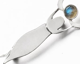 May include: A silver brooch featuring a stylised figure holding a round, iridescent gemstone.
