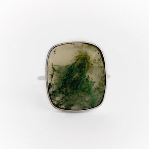May include: A silver ring with a square moss agate stone. The stone has a natural pattern of green and white. The ring is shown on a white background, highlighting the stone's details.