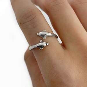 Puede incluir: Un anillo de plata con dos figuras de delfines que miran en direcciones opuestas. El anillo es ajustable y se puede llevar en cualquier dedo.